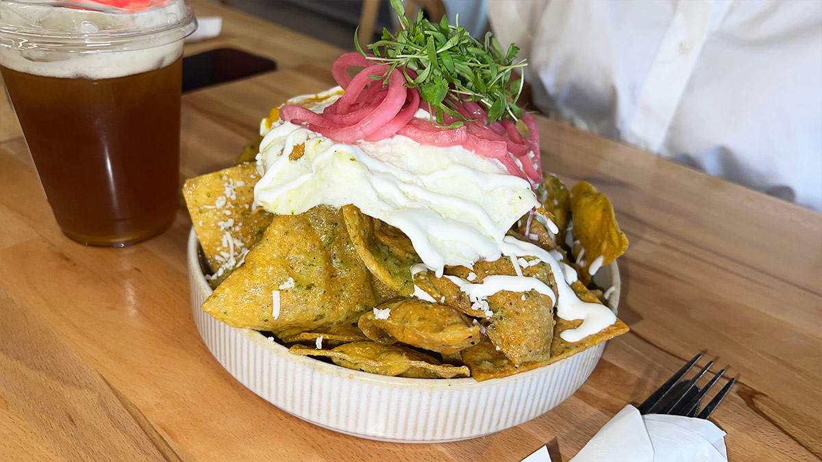 Chilaquiles in a bowl topped with a sunny-side-up egg, crema, pickled red onions, and herbs, with an iced tea nearby.