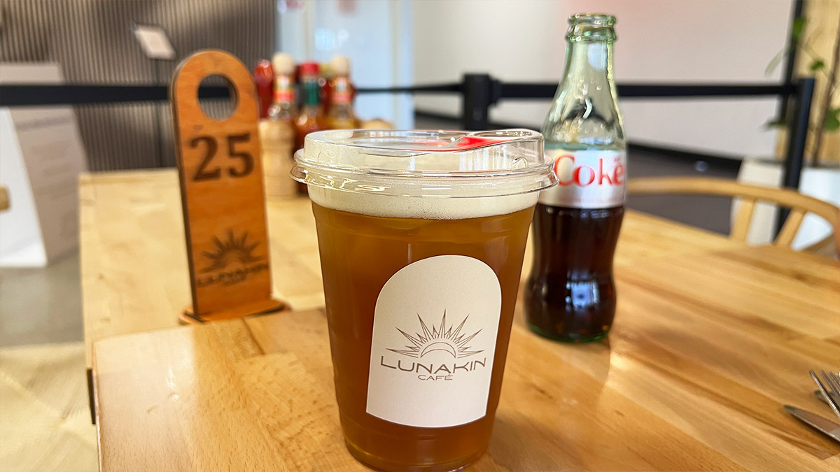 Plastic cup of iced tea with Lunakin Café logo on the table, with a glass bottle of Coke behind it.