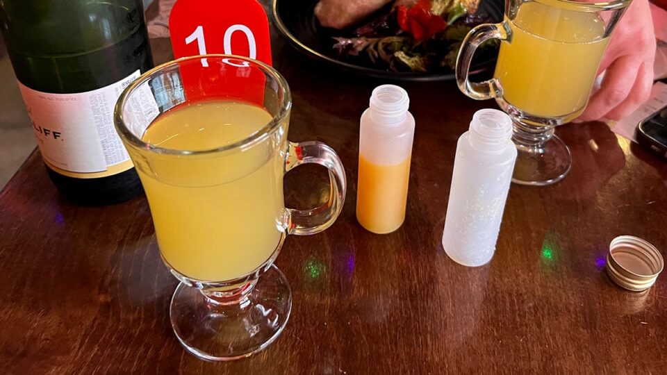 Two mimosa-style drinks in glass mugs beside a champagne bottle on a wooden table.