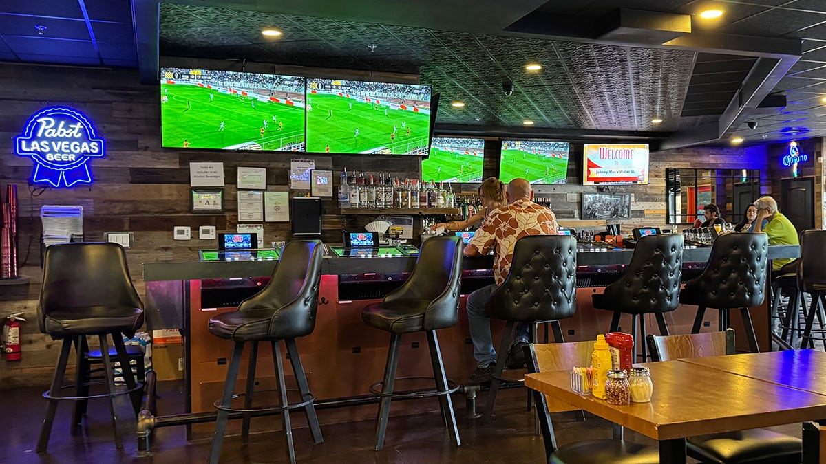 Interior of Johnny Mac’s bar featuring a video gaming machine and a TV showing a soccer match, with a lively and casual atmosphere.