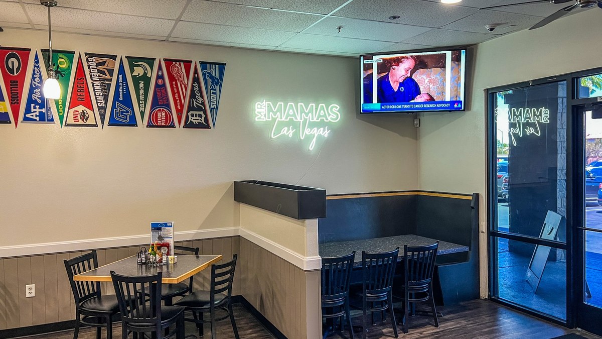 Dining room seating area inside Mr. Mamas Las Vegas with neon wall sign and sports pennants.