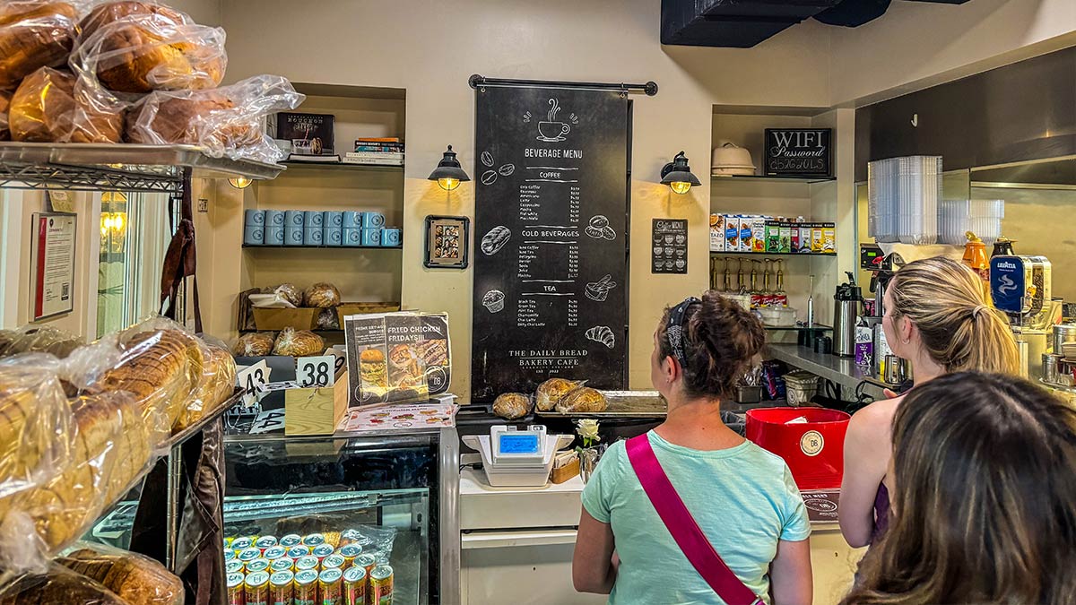 Customers standing at a bakery cafe counter with bread shelves and menu board in the background.