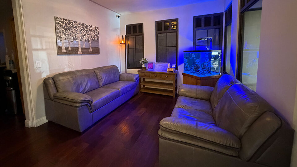 Spa waiting room with two gray leather sofas, wood flooring, framed artwork, and a blue-lit aquarium.