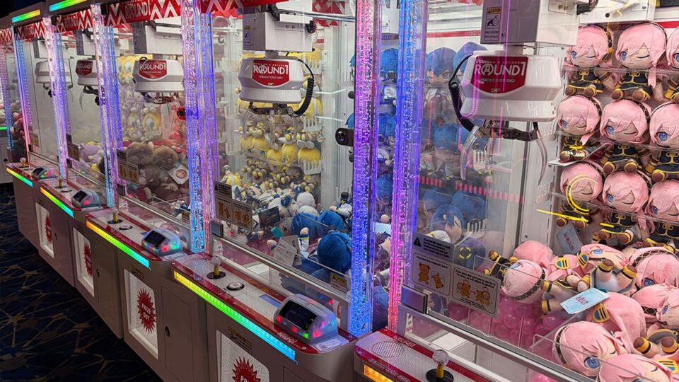 Row of illuminated Round1 claw machines filled with colorful anime character plush toys, including pink- and blue-haired dolls.