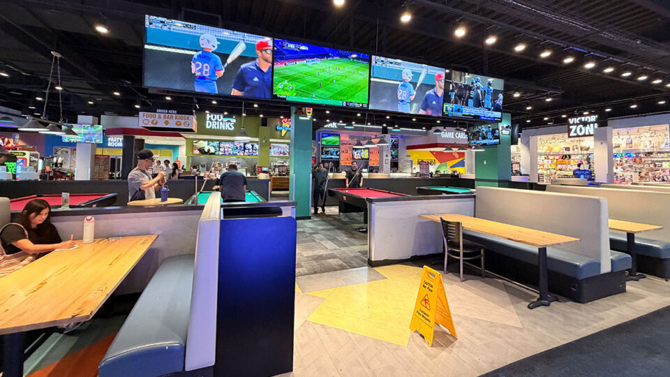 Dining booths and wooden tables beside billiards tables at Round1, with a food counter, overhead TVs, and prize store in the background.