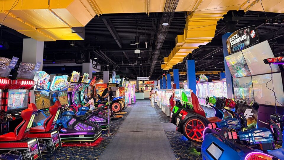 Wide view of Round1's arcade floor with colorful racing simulators, motorcycle-style games, and claw machines lining the walkway.