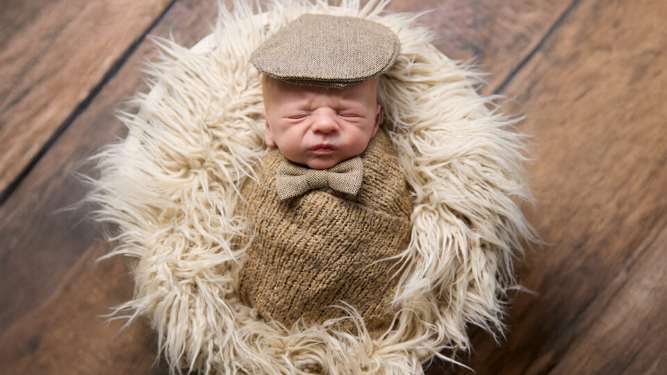 Newborn Sullivan wrapped in a brown knit blanket wearing a matching cap and bow tie